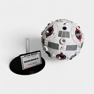 May include: A white and gray spherical training remote from Star Wars: Episode IV - A New Hope. The remote is labeled "MARKSMAN-H - Training Remote" and is on a black stand.