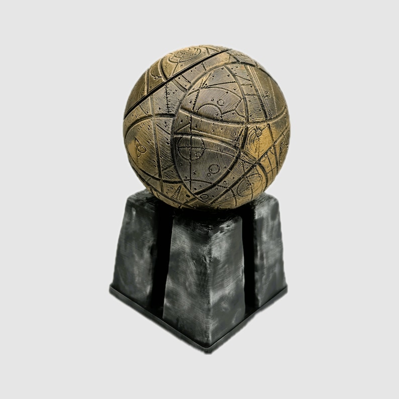 May include: A gold sphere with intricate patterns and markings sits on a black, square base. The sphere appears to be made of metal and has a textured surface.