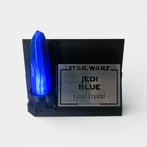 May include: A blue Kyber crystal replica from Star Wars, mounted on a black base. The base features a silver plaque with the text "STAR WARS JEDI BLUE - Kyber Crystal -". The crystal is a vibrant blue color and is the focal point of the display.