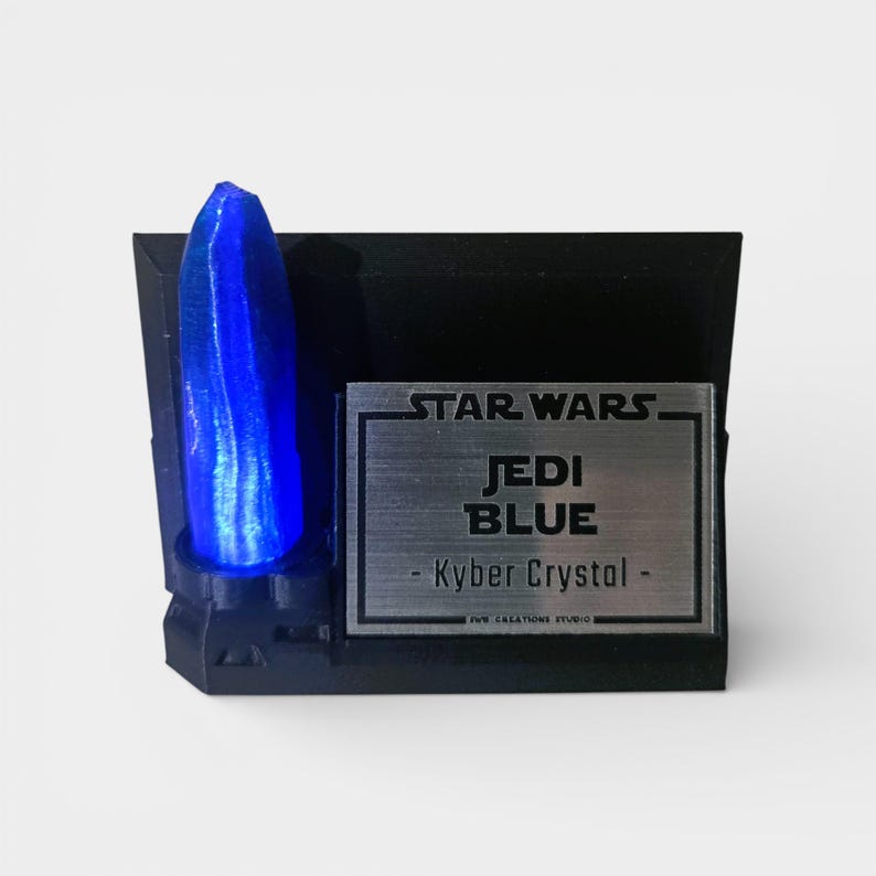 May include: A blue Kyber crystal replica from Star Wars, mounted on a black base. The base features a silver plaque with the text "STAR WARS JEDI BLUE - Kyber Crystal -". The crystal is a vibrant blue color and is the focal point of the display.