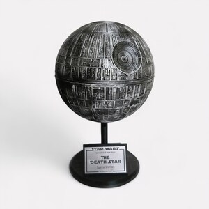Peut inclure: Maquette d&eacute;taill&eacute;e et m&eacute;tallique de l'&Eacute;toile de la Mort de Star Wars, &Eacute;pisode IV : Un nouvel espoir. La station spatiale sph&eacute;rique est mont&eacute;e sur un socle noir avec une plaque indiquant "STAR WARS - THE DEATH STAR - Space Station".