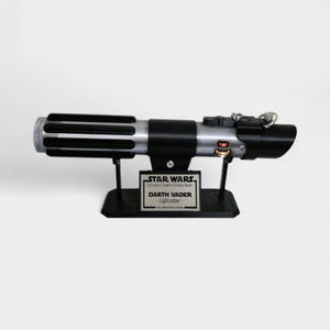 May include: A black and silver lightsaber replica, displayed on a black stand. The stand features a plaque with the text "STAR WARS Episode V Empire Strikes Back" and "DARTH VADER - Lightsaber-". The lightsaber has a detailed design.