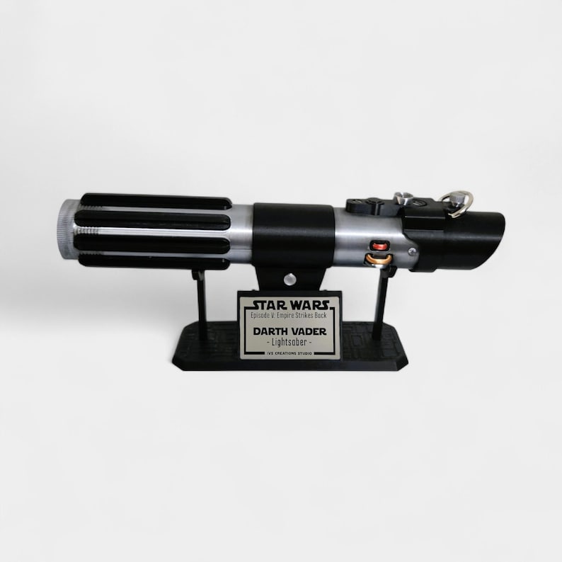 May include: A black and silver lightsaber replica, displayed on a black stand. The stand features a plaque with the text "STAR WARS Episode V Empire Strikes Back" and "DARTH VADER - Lightsaber-". The lightsaber has a detailed design.