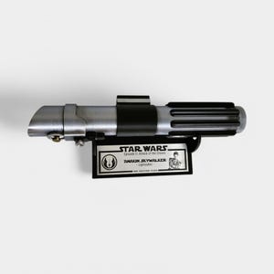 May include: A silver and black lightsaber replica from Star Wars: Episode II - Attack of the Clones. The replica is mounted on a black plaque with a silver inscription that reads "STAR WARS Episode II: Attack of the Clones ANAKIN SKYWALKER - Lightsaber -", and a small image of Anakin Skywalker.
