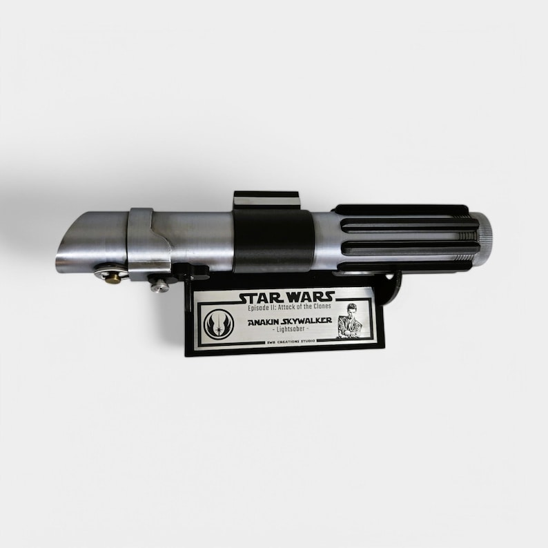 May include: A silver and black lightsaber replica from Star Wars: Episode II - Attack of the Clones. The replica is mounted on a black plaque with a silver inscription that reads "STAR WARS Episode II: Attack of the Clones ANAKIN SKYWALKER - Lightsaber -", and a small image of Anakin Skywalker.