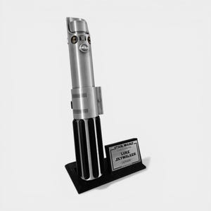 May include: A silver lightsaber replica, standing upright on a black base. The lightsaber has a detailed metallic finish with black accents. A small plaque next to it reads "STAR WARS" and "LUKE SKYWALKER - Lightsaber."