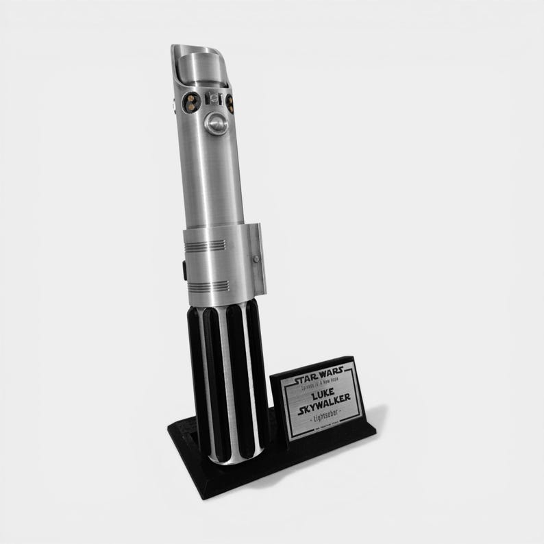 May include: A silver lightsaber replica, standing upright on a black base. The lightsaber has a detailed metallic finish with black accents. A small plaque next to it reads "STAR WARS" and "LUKE SKYWALKER - Lightsaber."