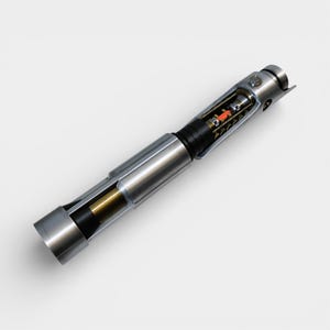 May include: A detailed, cross-section view of a metallic lightsaber prop. The silver hilt reveals internal components, including a red and black core. The design features a cylindrical shape with intricate details, showcasing a blend of mechanical and futuristic elements.
