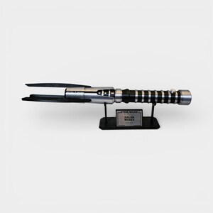 Op de afbeelding: Een zilveren en zwart lichtzwaard rekwisiet uit de Star Wars-franchise, met een zwarte handgreep en een zwart lemmet. De rekwisiet wordt tentoongesteld op een zwarte standaard met een naamplaatje met de tekst "Star Wars Galen Marek Lightsaber".