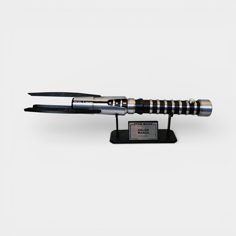 Op de afbeelding: Een zilveren en zwart lichtzwaard rekwisiet uit de Star Wars-franchise, met een zwarte handgreep en een zwart lemmet. De rekwisiet wordt tentoongesteld op een zwarte standaard met een naamplaatje met de tekst "Star Wars Galen Marek Lightsaber".