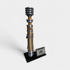 Puede incluir: R&eacute;plica de sable de luz met&aacute;lico con detalles plateados, dorados y negros. El sable de luz se exhibe sobre un soporte negro con una peque&ntilde;a placa que dice "STAR WARS LEIA ORGANA Lightsaber."