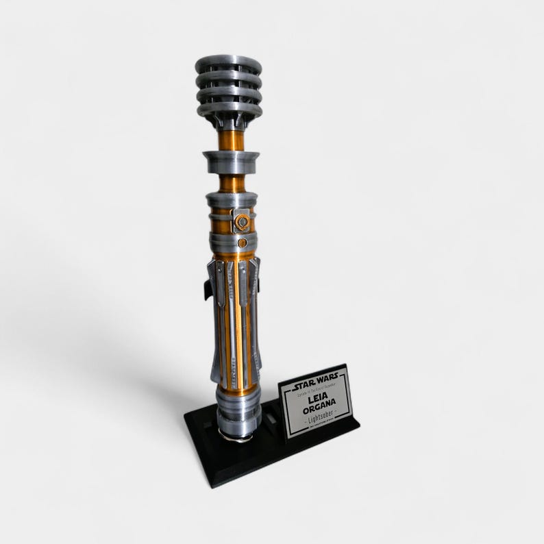 Puede incluir: R&eacute;plica de sable de luz met&aacute;lico con detalles plateados, dorados y negros. El sable de luz se exhibe sobre un soporte negro con una peque&ntilde;a placa que dice "STAR WARS LEIA ORGANA Lightsaber."