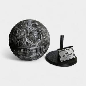Peut inclure: Maquette sph&eacute;rique d&eacute;taill&eacute;e de l'&Eacute;toile de la Mort de Star Wars, dans des tons de gris et de noir. Le mod&egrave;le est &agrave; c&ocirc;t&eacute; d'un socle noir avec une plaque indiquant "STAR WARS The Death Star - Space Station -."