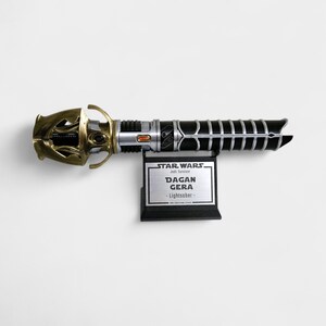 May include: A black and silver lightsaber with a gold handle. The lightsaber is on a black stand with a silver plaque that reads "Star Wars Jedi: Survivor Dagan Gera - Lightsaber - The Creature Cantina".