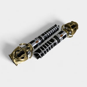 May include: Two black and gold lightsaber hilts with intricate details. The hilts are made of metal and have a textured surface. The hilts are separated and lying on a white background.
