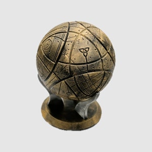 May include: A gold-colored sphere with intricate patterns and symbols, resting on a matching stand. The sphere has a textured surface and appears to be made of metal.