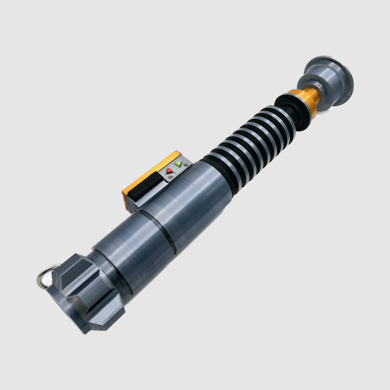 May include: A gray and gold lightsaber toy with a black and silver handle and a black and silver blade. The lightsaber has a small, black, rectangular control panel with a green and red light.