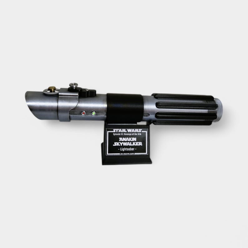 May include: A silver and black lightsaber with a black handle and a silver blade. The lightsaber is mounted on a black stand with a plaque that reads "STAR WARS Episode III: Revenge of the Sith ANAKIN SKYWALKER - Lightsaber -"