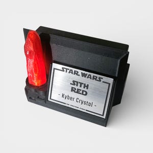 May include: A black display stand featuring a vibrant red Kyber crystal. The stand has a silver plaque with "STAR WARS SITH RED - Kyber Crystal" text. The design is angular and modern, with a focus on the crystal's color.
