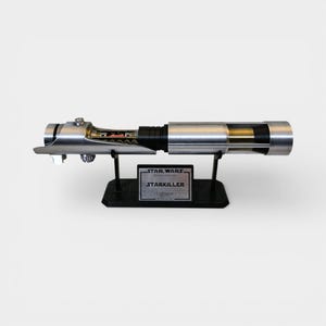 May include: A silver and black lightsaber replica, displayed on a black stand. The lightsaber features intricate detailing and a clear section revealing internal components. The stand includes a plaque that reads "STAR WARS STARKILLER lightsaber".
