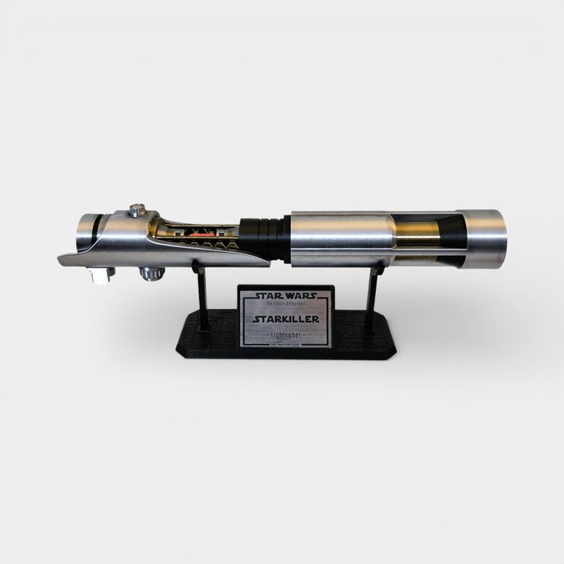 May include: A silver and black lightsaber replica, displayed on a black stand. The lightsaber features intricate detailing and a clear section revealing internal components. The stand includes a plaque that reads "STAR WARS STARKILLER lightsaber".