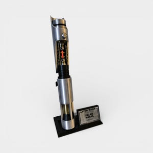 May include: A silver and black lightsaber prop with a detailed internal view, featuring a red core. The prop is displayed on a black stand with a plaque that reads "STAR WARS, GALEN MAREK, Lightsaber."