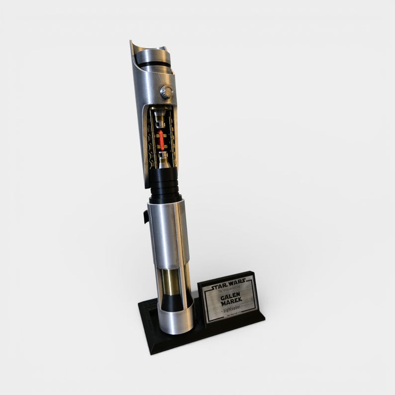 May include: A silver and black lightsaber prop with a detailed internal view, featuring a red core. The prop is displayed on a black stand with a plaque that reads "STAR WARS, GALEN MAREK, Lightsaber."