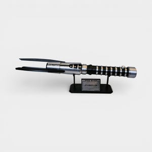 Op de afbeelding: Een zilver-zwarte lichtzwaard replica, gepresenteerd op een zwarte standaard. Het lichtzwaard heeft een gedetailleerde handgreep met zwarte en zilveren accenten. De standaard heeft een plaatje met de tekst "STAR WARS STARKILLER".