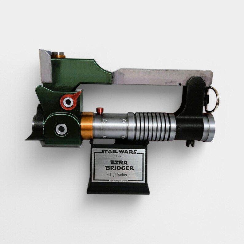 Może przedstawiać: Replika miecza świetlnego Star Wars w kolorze zielonym, czarnym i srebrnym z tabliczką z napisem "Star Wars Ezra Bridger - Miecz świetlny - The Mandalorian"