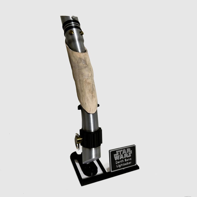 May include: A silver lightsaber with a brown leather wrap on the handle. The lightsaber is on a black stand with a silver plaque that reads "Star Wars Darth Bane Lightsaber".