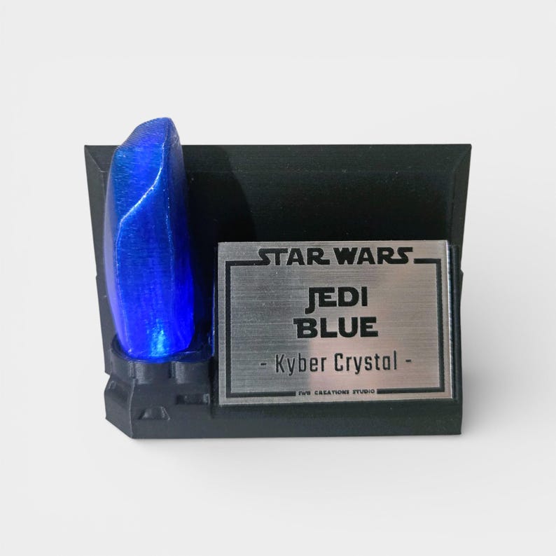 May include: A blue Kyber Crystal replica from Star Wars, mounted on a black base. The base features a silver plaque with the text "STAR WARS JEDI BLUE - Kyber Crystal - ". The crystal is a vibrant blue color.