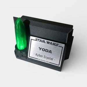 May include: A black Star Wars display featuring a green Kyber Crystal and a silver nameplate with "YODA" and "Kyber Crystal" text. The display has a textured design and is set against a light background.