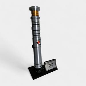 May include: A silver and black lightsaber with a red button and gold accents, displayed on a black stand. The stand features a small plaque with the text "STAR WARS Darth Revan Lightsaber".