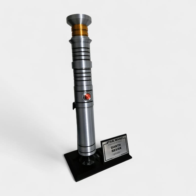 May include: A silver and black lightsaber with a red button and gold accents, displayed on a black stand. The stand features a small plaque with the text "STAR WARS Darth Revan Lightsaber".