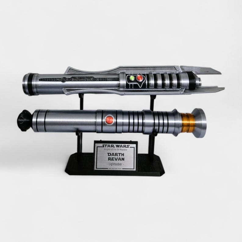 May include: Two silver lightsabers, one above the other, displayed on black stands. The top saber has a unique design with red and green accents. The bottom saber has a red button and gold-colored details. A plaque reads "STAR WARS DARTH REVAN - Lightsaber -."