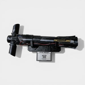 May include: A black Kylo Ren lightsaber from Star Wars: The Force Awakens, with a crossguard design. The lightsaber is mounted on a black base with a nameplate that reads "Kylo Ren - Lightsaber".