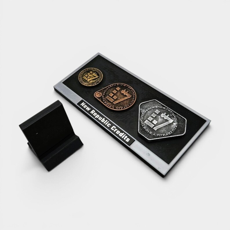 May include: A black rectangular display featuring three New Republic Credits coins in gold, bronze, and silver. The coins have a geometric design. The display also includes a small black stand. The text "New Republic Credits" is visible.