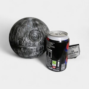 Peut inclure: Une maquette d&eacute;taill&eacute;e et m&eacute;tallique de l'&Eacute;toile de la Mort &agrave; c&ocirc;t&eacute; d'une canette de soda noire avec des accents rouges et bleus. La canette porte l'inscription "STAR WARS - The Death Star - Space Station".