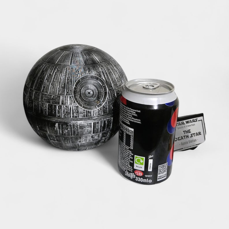 Peut inclure: Une maquette d&eacute;taill&eacute;e et m&eacute;tallique de l'&Eacute;toile de la Mort &agrave; c&ocirc;t&eacute; d'une canette de soda noire avec des accents rouges et bleus. La canette porte l'inscription "STAR WARS - The Death Star - Space Station".