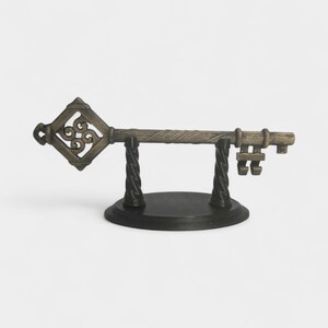 May include: An antique-style decorative key, displayed on a black stand. The key is bronze-colored with intricate detailing, including a diamond-shaped head with a scrolled design. The stand has a round base and two twisted supports.