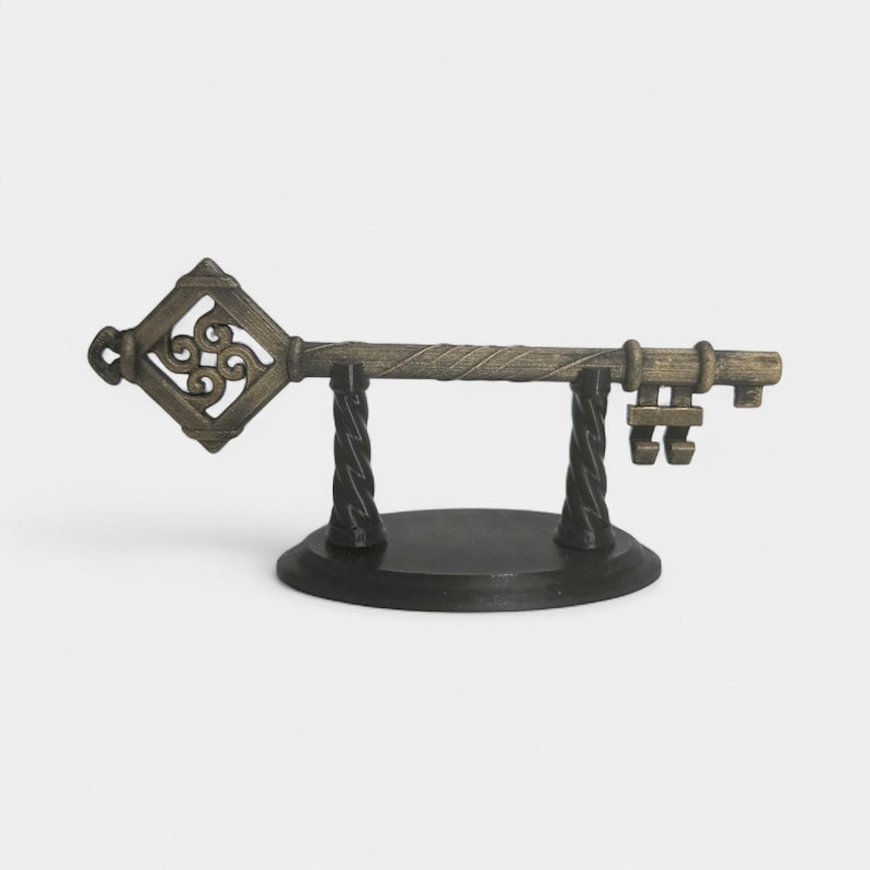 May include: An antique-style decorative key, displayed on a black stand. The key is bronze-colored with intricate detailing, including a diamond-shaped head with a scrolled design. The stand has a round base and two twisted supports.
