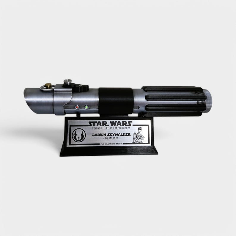 May include: A silver and black lightsaber replica from Star Wars: Episode II - Attack of the Clones. The lightsaber is on a black display stand with a silver plaque that reads "STAR WARS Episode II: Attack of the Clones ANAKIN SKYWALKER - Lightsaber - FXW CREATIONS STUDIO".