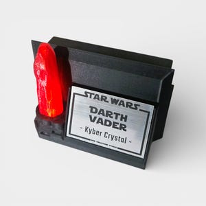 May include: A black display stand featuring a red Kyber Crystal, with the text "STAR WARS DARTH VADER - Kyber Crystal" on a silver plaque. The stand is made of black material and has a rectangular shape.