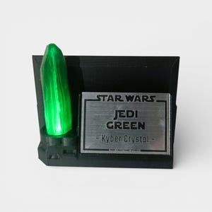 May include: A black display stand featuring a vibrant green Kyber Crystal, labeled "Jedi Green" and "Star Wars." The stand has a metallic nameplate with the text "Kyber Crystal."