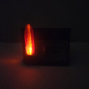 May include: A glowing, orange-red crystal, possibly a prop, is set in a dark, rectangular base. The crystal emits a warm light, with the base appearing to have text. The overall effect is dramatic and mysterious.