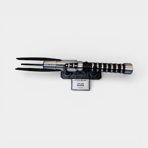 Op de afbeelding: Een zilveren en zwarte lichtzwaard met een drietandig blad, gemonteerd op een zwarte basis. De basis heeft het Star Wars-logo en een label met de tekst "GALEN MAREK - Lightsaber".