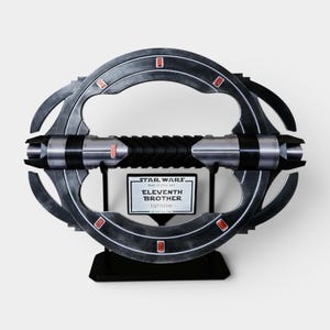 K&ouml;nnte beinhalten: Ein Star Wars Eleventh Brother Lichtschwert-Requisite. Das Lichtschwert ist silber und schwarz mit roten Akzenten. Der Griff ist schwarz mit gerippter Textur. Das Lichtschwert wird auf einem schwarzen St&auml;nder mit einem Namensschild ausgestellt.