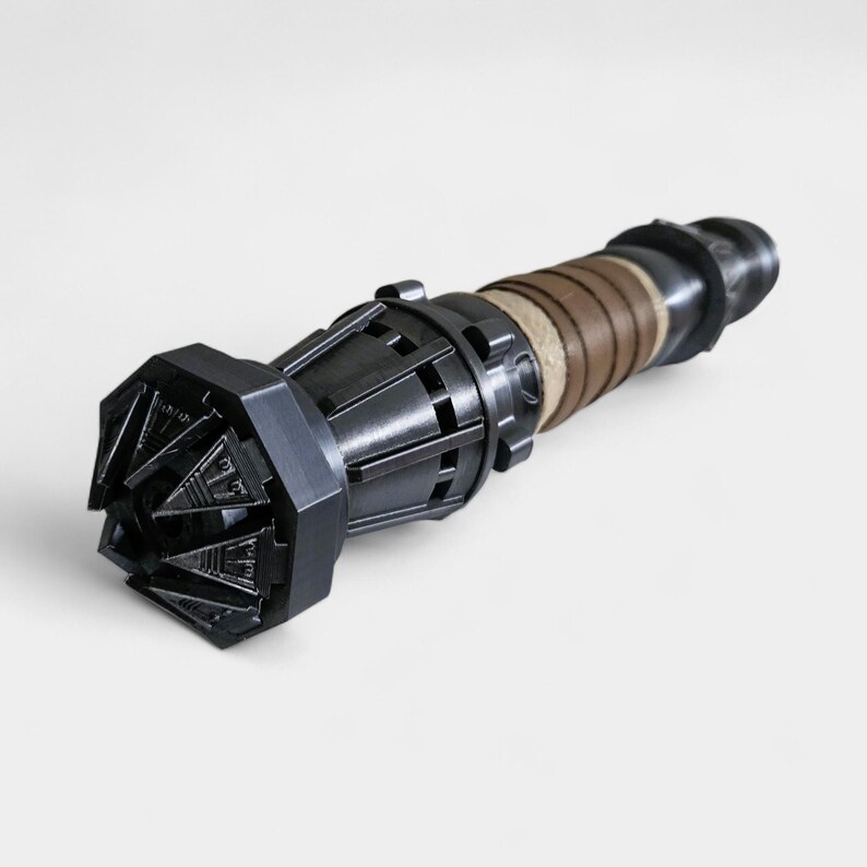 May include: A black and brown lightsaber with a detailed, intricate design. The lightsaber has a textured, cylindrical handle and a pointed, multi-faceted blade.