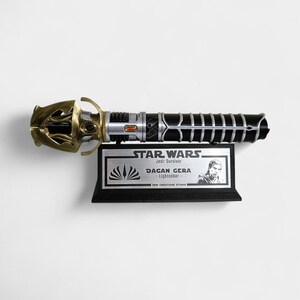 May include: A black and silver lightsaber with a gold hilt, displayed on a black plaque with the text "STAR WARS Jedi: Survivor DAGAN GERA - Lightsaber - THE CREATIONS STUDIO".