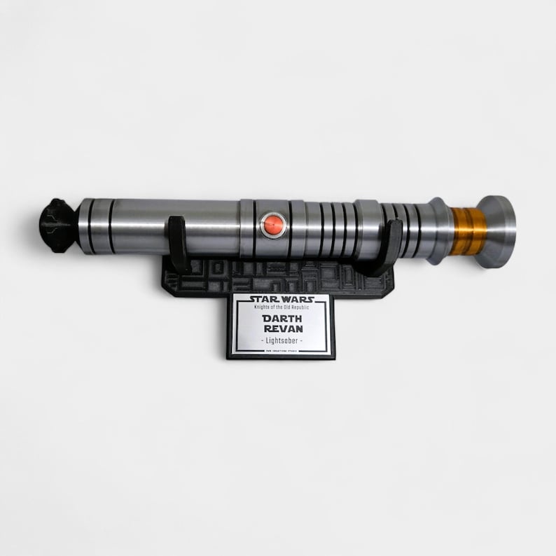 May include: A silver and black Darth Revan lightsaber replica, mounted on a black display stand. The lightsaber features a red button and gold accents. The stand includes the Star Wars logo and the text "Darth Revan - Lightsaber."